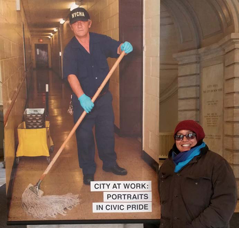 NYCHA photographer Leticia Barboza stands with her portrait of Caretaker Pellumb Skenderi in the “City at Work: Portraits in Civic Pride” exhibit in City Hall.