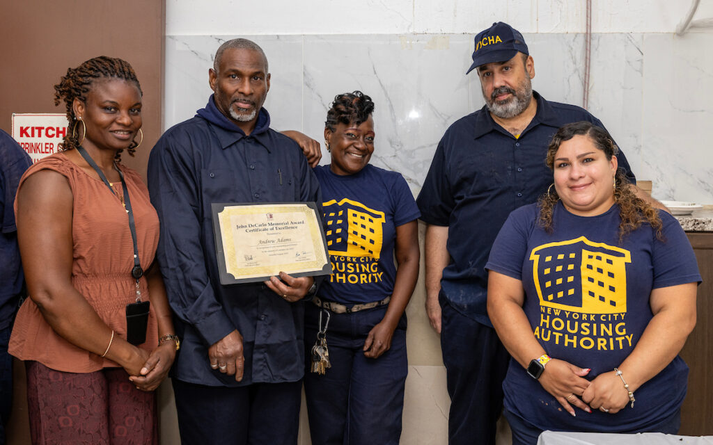 A group of five individuals, including one holding a certificate, stand together in a room with a “New York City Housing Authority” logo on their shirts.