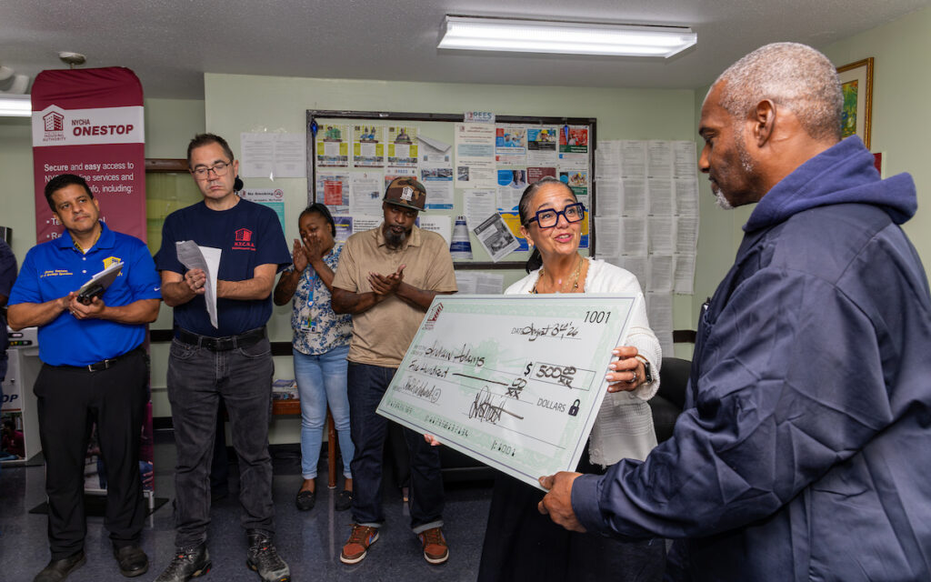 A person receives a large ceremonial check for $1,500 from another individual in a community center setting, with others observing and applauding.