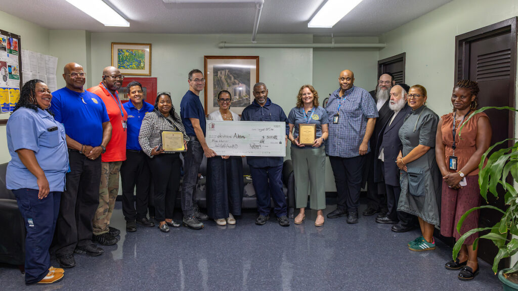 A group of people in an office setting pose with a large ceremonial check and framed certificates, smiling for the photo.