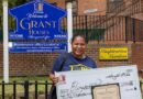 A person stands outdoors holding a large ceremonial check for $1,000 and an award certificate, in front of a NYCHA sign for Grant Houses.