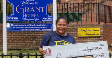 A person stands outdoors holding a large ceremonial check for $1,000 and an award certificate, in front of a NYCHA sign for Grant Houses.