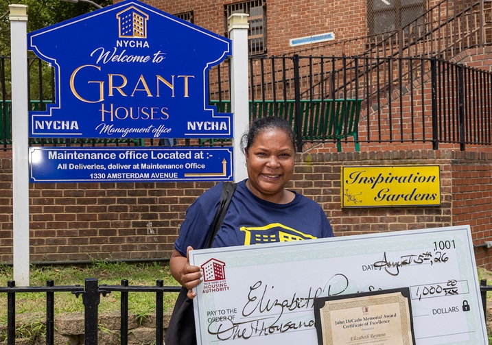 A person stands outdoors holding a large ceremonial check for $1,000 and an award certificate, in front of a NYCHA sign for Grant Houses.
