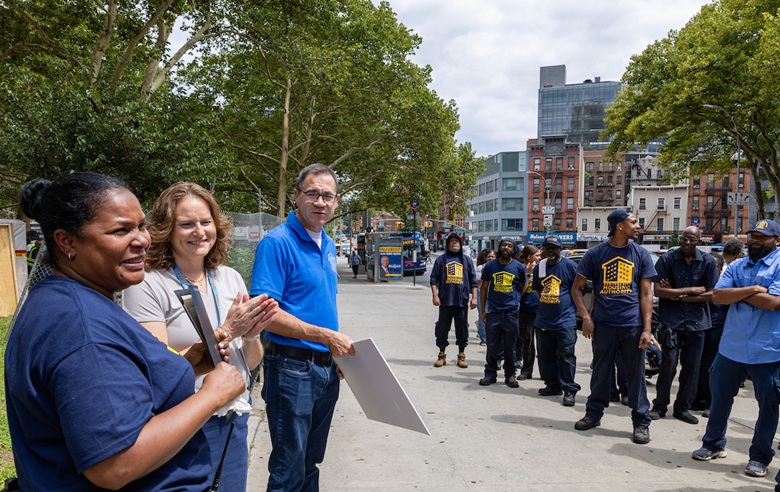 A group of people, including workers in matching shirts, gather on a city sidewalk for what appears to be a public meeting or demonstration.