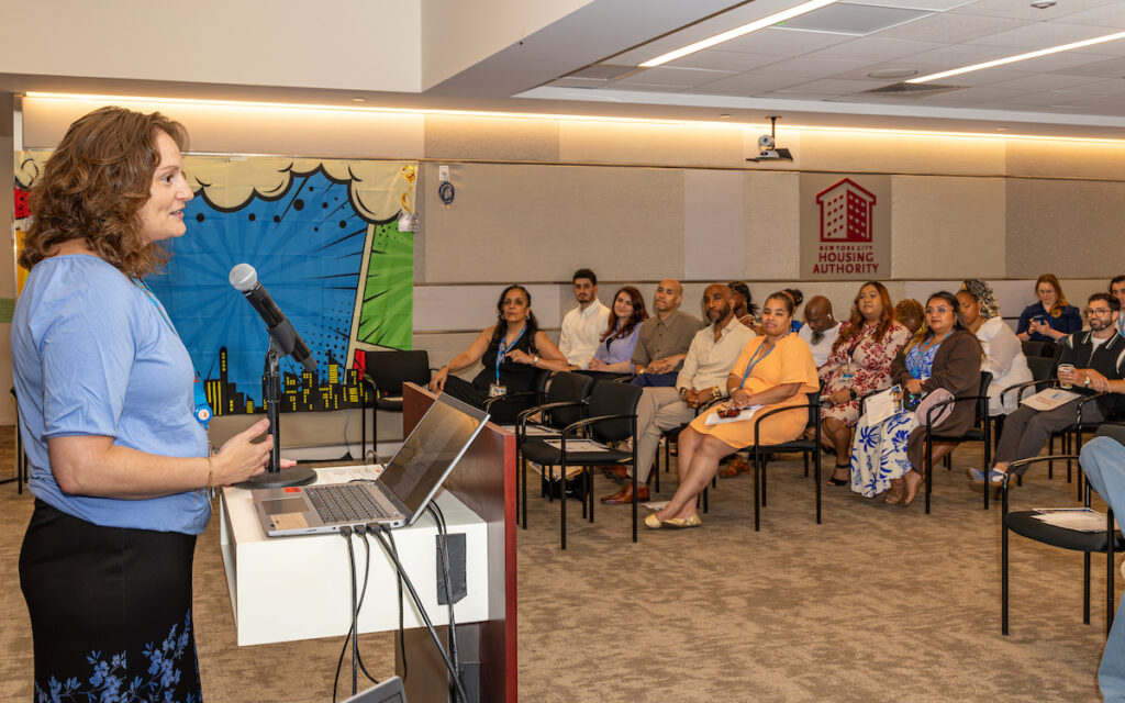 A person speaks at a podium in front of an audience seated in a conference room, with a sign for the New York City Housing Authority visible on the wall.