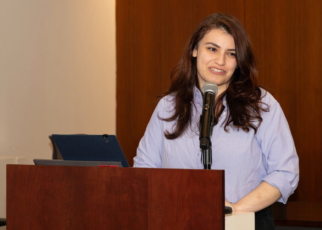 A person with long dark hair stands at a wooden podium, speaking into a microphone while wearing a light blue shirt.