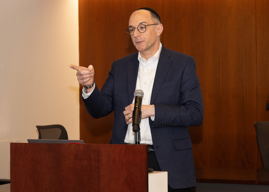 A person in a dark suit and glasses stands at a podium, gesturing while speaking into a microphone.