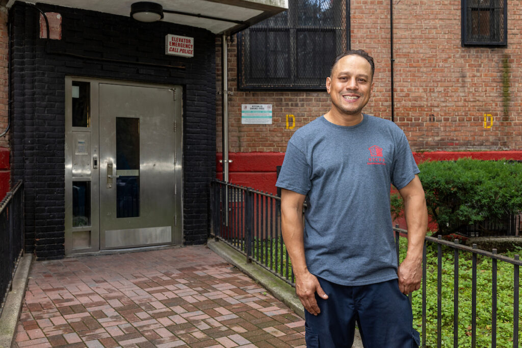 A person stands smiling in front of a brick building entrance with a metal door and an emergency call sign.
