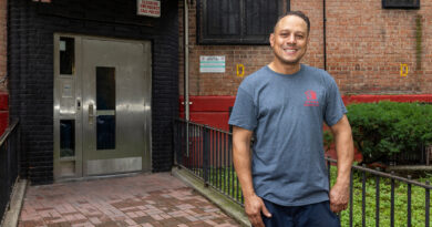 A person stands smiling in front of a brick building entrance with a metal door and an emergency call sign.