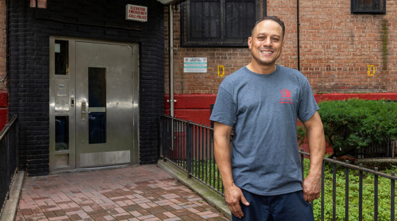 A person stands smiling in front of a brick building entrance with a metal door and an emergency call sign.