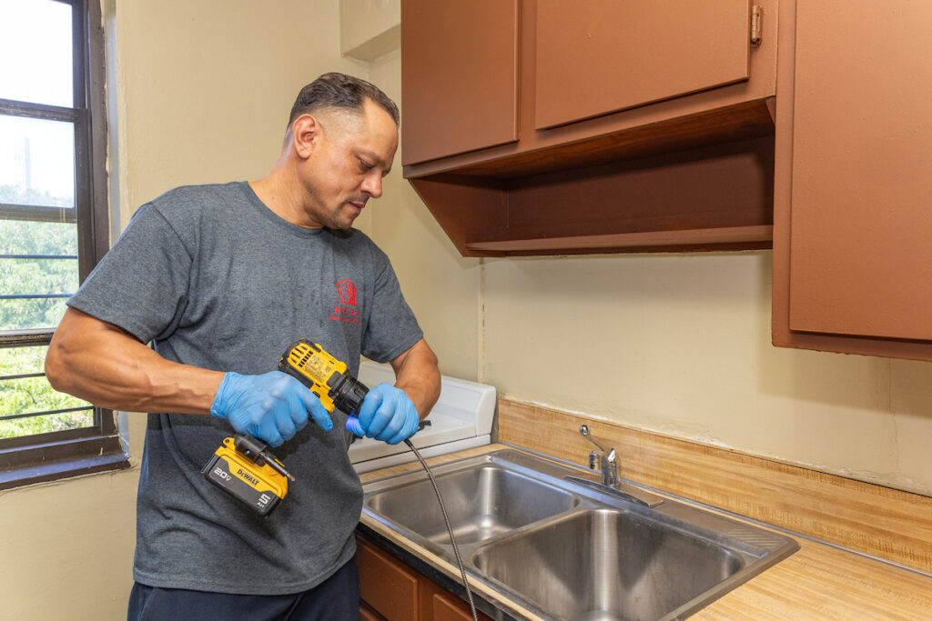 A person wearing blue gloves uses a yellow cordless drill to work on a kitchen sink.