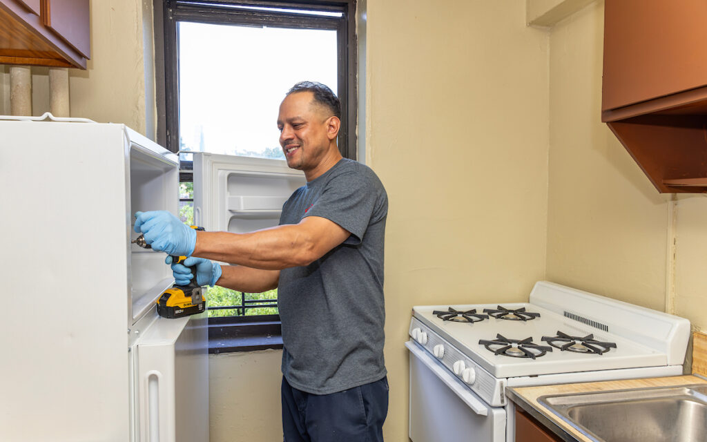 A person wearing blue gloves uses a power drill to work on the inside of a white refrigerator in a kitchen.