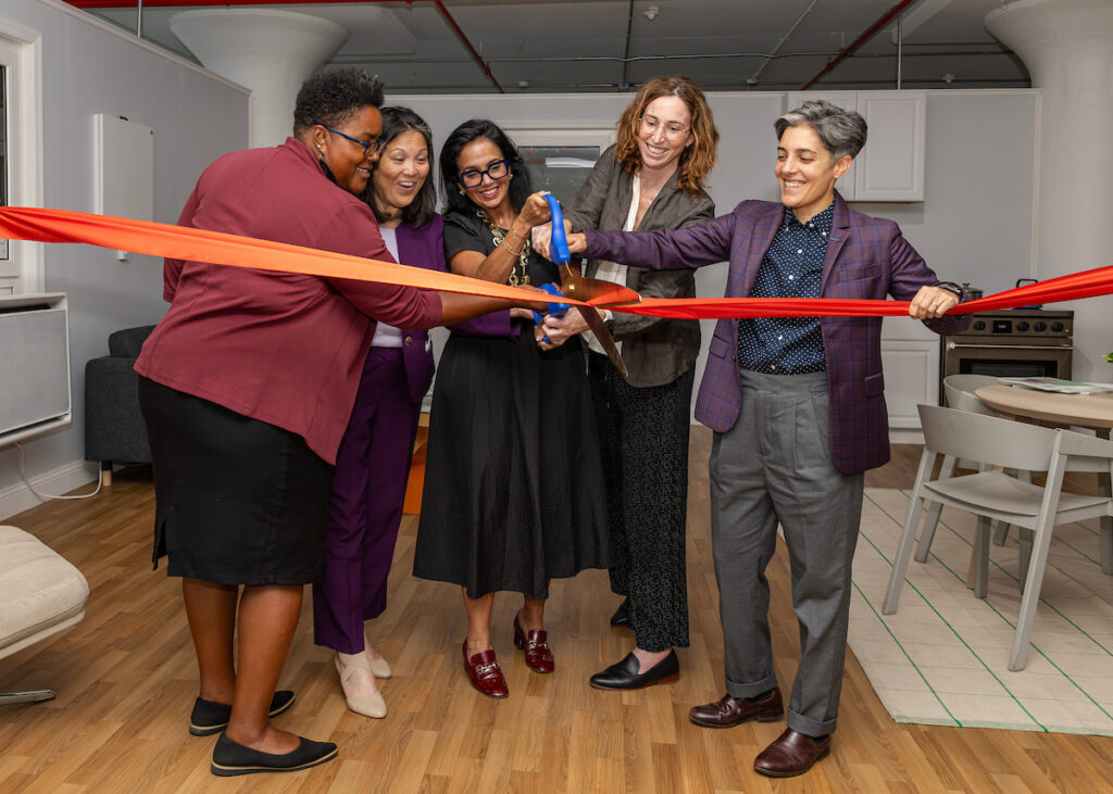 Five individuals joyfully cut a vibrant red ribbon together in a modern, brightly lit indoor space.